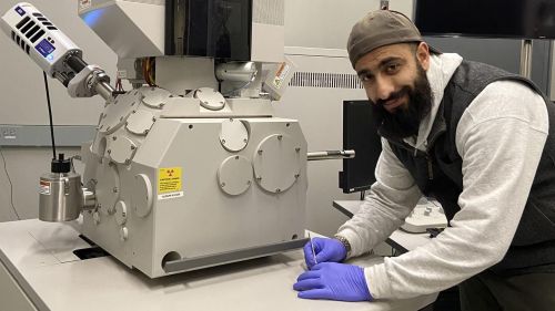 Male student wearing latex gloves, smiling in front of imaging equipment in lab.