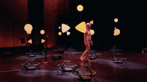 In a dimly lit theater, Naomi Leonard walks among a group of tall, slender robots, each spotlit and decorated with colorful cutouts of circles and triangles.