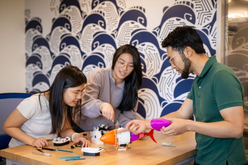 Three people work together on assembling robotic fish, with tools and electronic parts spread on the table.