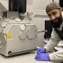 Male student wearing latex gloves, smiling in front of imaging equipment in lab.