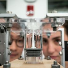 Close-up of a glass cylinder containing a thick, transparent substance with a bubbly pattern forming on the top layer, and two students’ faces peering closely at the experiment.