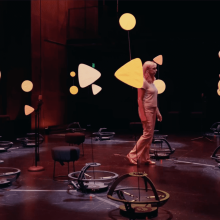 In a dimly lit theater, Naomi Leonard walks among a group of tall, slender robots, each spotlit and decorated with colorful cutouts of circles and triangles.
