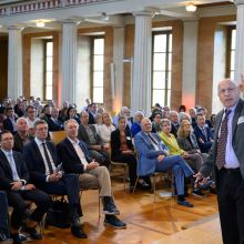 Howard Stone speaks on stage to a large seated audience in a hall with tall white columns and large windows